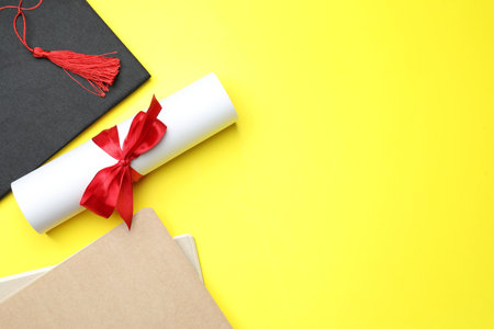 Diploma, graduation hat and notebooks on yellow background, flat lay. Space for textの写真素材