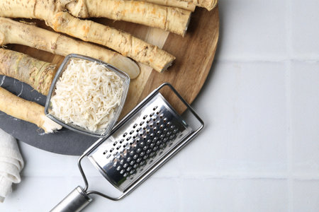 Whole and gratified horseradish roots near grater on white tiled table, top view. Space for textの写真素材