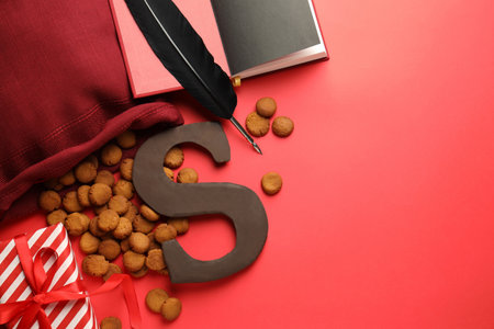 Saint Nicholas (Sinterklaas) day. Book, feather, chocolate letter S, kruidnoten cookies and gift box on red background, flat lay. Space for textの写真素材