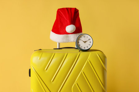 Stylish suitcase with Santa hat and alarm clock on yellow background, closeup. Christmas vacationの写真素材
