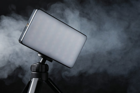 LED lamp on tripod against black background with smoke, closeup. Space for textの写真素材