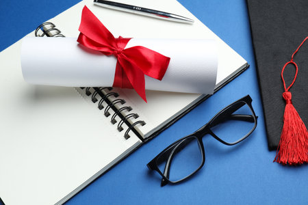 Diploma, graduation hat, notebook, glasses and pen on blue backgroundの写真素材