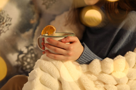 Cozy winter evening. Woman wrapped in plaid enjoying hot drink near window, closeup. Bokeh effectの写真素材