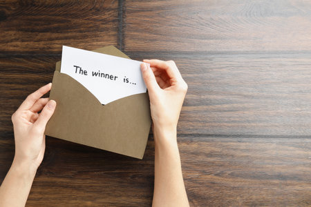 Woman taking card with text The Winner Is from envelope at wooden table, top view. Space for textの写真素材