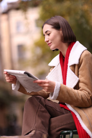 Beautiful woman in coat reading newspaper on chair outdoorsの写真素材