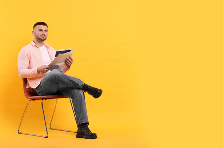 Young man reading newspaper on chair against yellow background. Space for textの写真素材