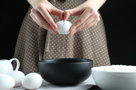 Woman cracking egg into bowl at table against black background, closeupの写真素材