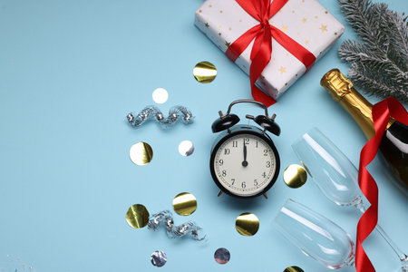 Alarm clock, bottle of sparkling wine, festive decor and gift box on light blue background, flat lay...