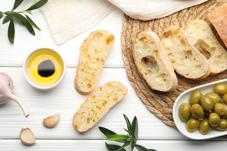 Fresh cut ciabatta, oil, olives, garlic and leaves on white wooden table, flat layの写真素材