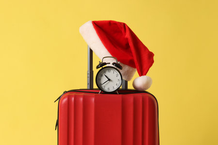 Stylish suitcase with Santa hat and alarm clock on yellow background, closeup. Christmas vacationの写真素材