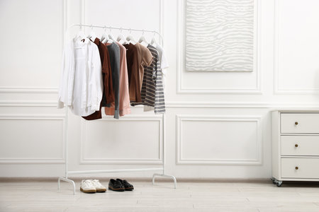 Different women's clothes hanging on rack and chest of drawers near white wall indoorsの写真素材