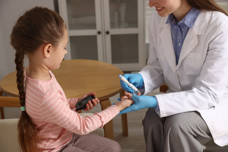 Diabetes. Smiling doctor in medical gloves checking little girl's blood sugar level with lancet pen and glucometer indoors, closeupの写真素材