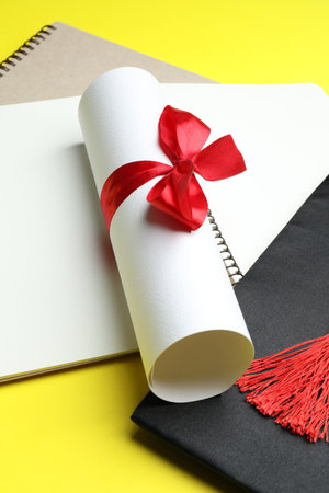 Diploma, graduation hat and notebooks on yellow background, closeupの写真素材
