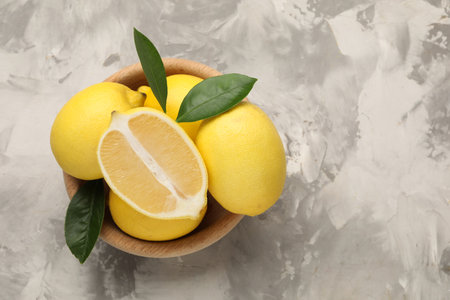 Whole and cut lemons with green leaves in bowl on gray textured table, top view. Space for text