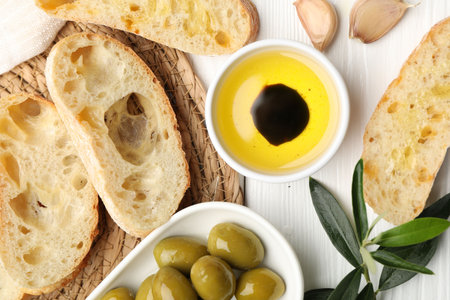 Fresh cut ciabatta, oil, olives, garlic and leaves on white wooden table, flat layの写真素材