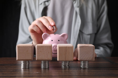Woman putting coin into piggy bank at wooden table, focus on blank cubes and money. Mockup for designの写真素材