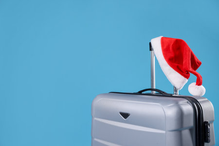 Christmas vacation. Suitcase with Santa hat on light blue background, closeup. Space for textの写真素材