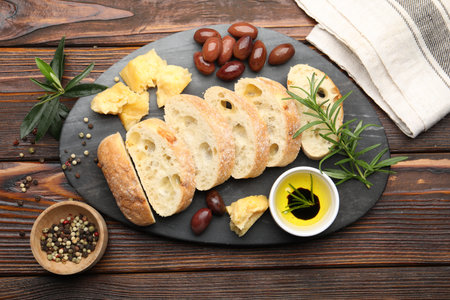 Pieces of fresh ciabatta, butter, olives, cheese, rosemary and peppercorns on wooden table, flat lay