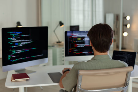 Programmer working on computer and laptop at white table in office, back view