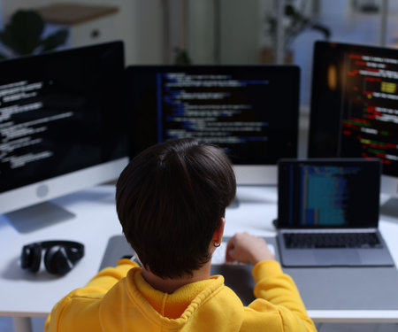 Programmer working on computer with triple monitors and laptop at white table in office, back viewの写真素材