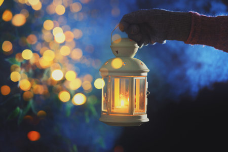 Woman holding lantern with candle against blurred lights on greeting cardの写真素材