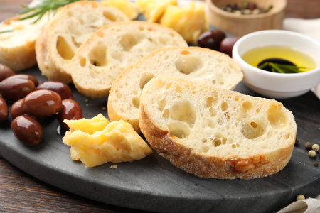 Pieces of fresh ciabatta, butter, olives, cheese and peppercorns on wooden table, closeup