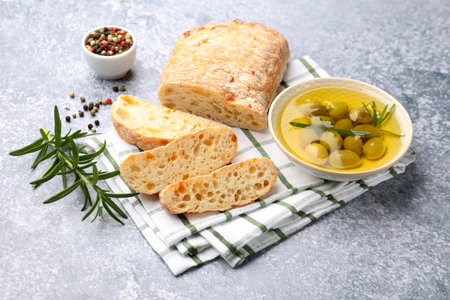 Fresh cut ciabatta, oil, olives, rosemary and peppercorns on gray textured table, closeup