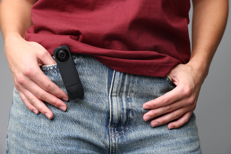 Young woman with hidden spy camera in her pocket on light gray background, closeup