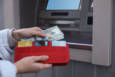 Woman putting money into wallet near cash machine outdoors, closeup. ATM