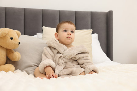 Cute baby in bathrobe and toy bear on bed at homeの写真素材
