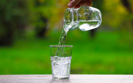 Man pouring water into glass from jug at wooden table against blurred green background, closeup....