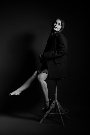 Beautiful young woman in stylish clothes sitting on chair against dark background. Black and white effectの写真素材