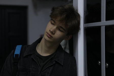 Depressed teenage boy with backpack near lockers indoors
