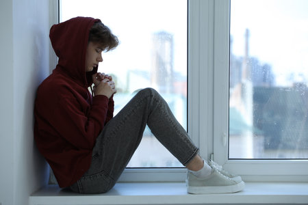 Depressed teenage boy near window at home
