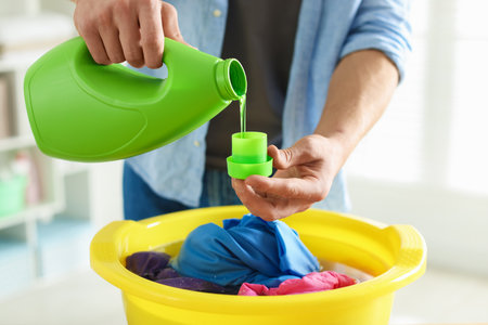 Man pouring detergent into cap over plastic basin with clothes indoors, closeupの写真素材