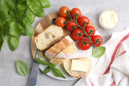 Slices of baguette with butter, basil, tomatoes and knife on light gray table, flat lay