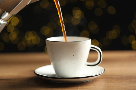 Pouring aromatic coffee into cup at wooden table against black background with blurred lights,...