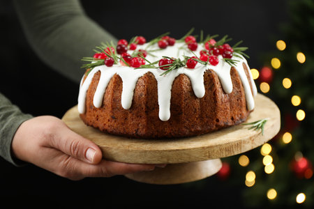 Woman holding delicious Christmas cake with icing, cranberries and rosemary against black background...