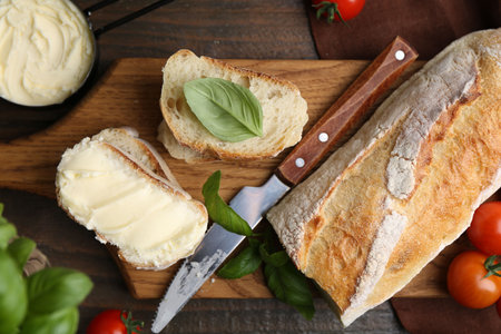 Slices of baguette with butter, basil, tomatoes and knife on wooden table, flat layの写真素材