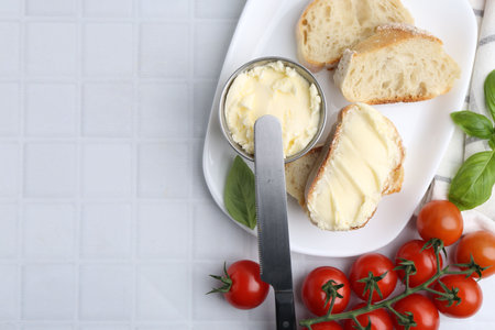 Slices of baguette with butter, basil, tomatoes and knife on white tiled table, flat lay. Space for textの写真素材