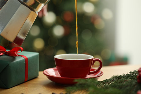 Pouring aromatic coffee from moka pot into cup at wooden table with gift box and fir tree branches against blurred lights, closeup. Christmas greeting cardの写真素材