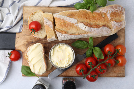 Cut baguette with butter, basil, tomatoes and knife on light table, flat layの写真素材