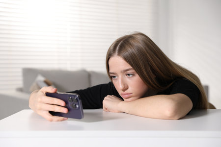 Sad teenage girl with smartphone at white table indoorsの写真素材