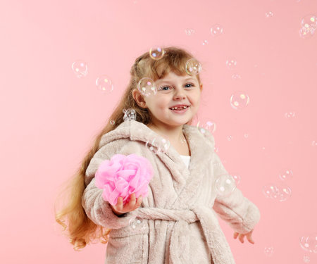 Cute girl in bathrobe with loofah and bubbles on pink backgroundの写真素材