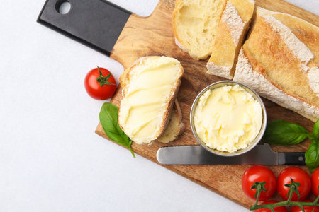 Cut baguette with butter, basil, tomatoes and knife on light table, flat lay. Space for textの写真素材