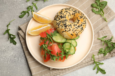 Delicious bagel with salmon, cucumber, arugula and lemon on gray table, flat lay