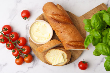 Cut baguette with butter, basil and tomatoes on white marble table, flat layの写真素材