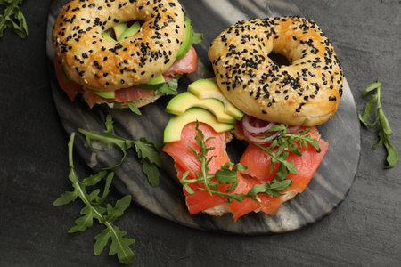Delicious bagels with salmon, avocado, onion and arugula on black table, flat layの写真素材