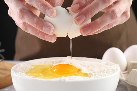 Woman breaking egg into bowl with flour at table against black background, closeupの写真素材