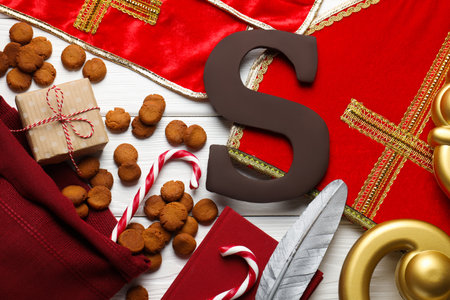 Sinterklaas (Saint Nicholas) Day. Chocolate letter S, kruidnoten, candy canes, mitre, stole, crosier and notebook with feather on white wooden table, flat layの写真素材